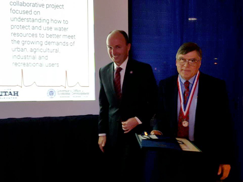 Two men stand and smile at the camera. On the left is Lieutenant Governor Spencer Cox, wearing a suit. On the right is Joel Harris, wearing a suit and his new Governor's Medal around his neck.